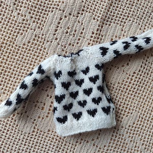 May include: A white knitted sweater with a black heart pattern. The sweater has long sleeves and a crew neck.
