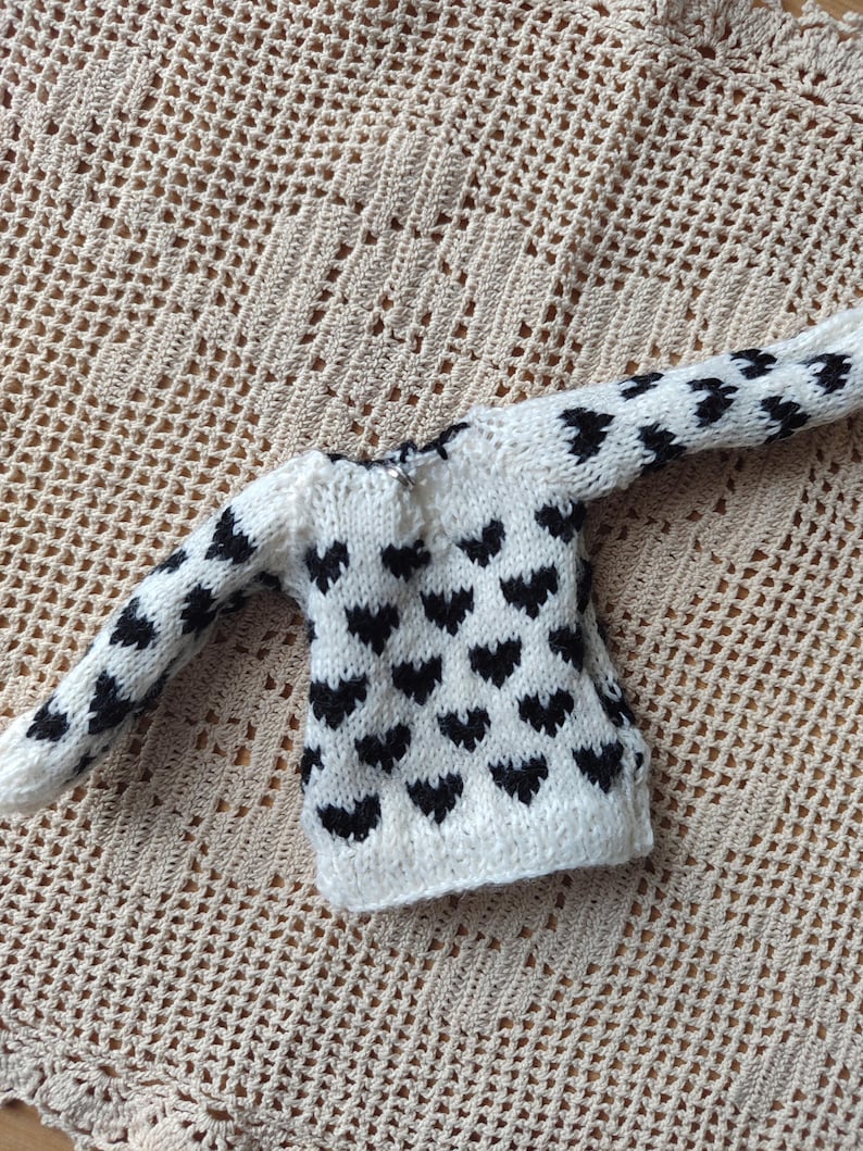 May include: A white knitted sweater with a black heart pattern. The sweater has long sleeves and a crew neck.