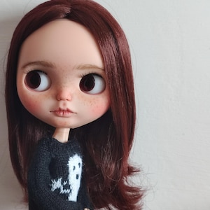 May include: A doll with long, dark brown hair wearing a black sweater with a white ghost design. The doll has large, brown eyes and a neutral expression.