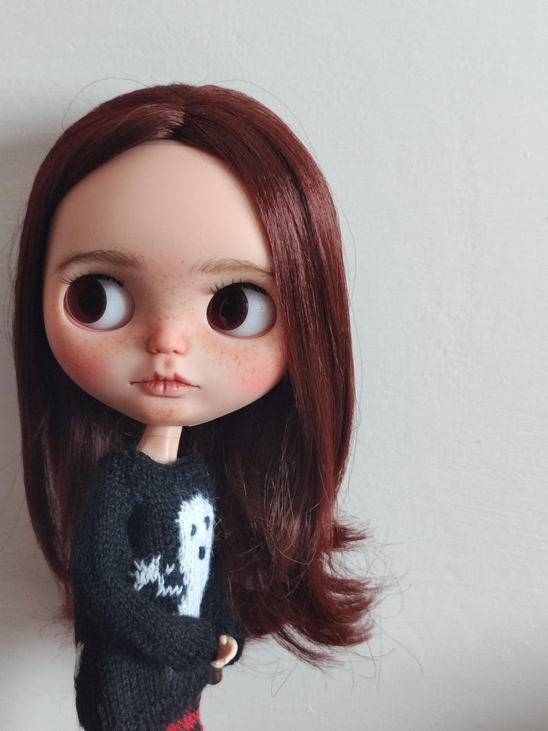 May include: A doll with long, dark brown hair wearing a black sweater with a white ghost design. The doll has large, brown eyes and a neutral expression.