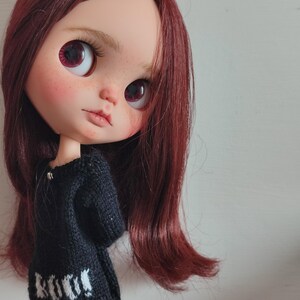 May include: A doll with long, dark red hair wearing a black sweater with white lettering that says "Knit" and red and black plaid pants.