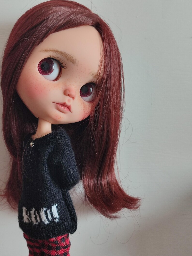 May include: A doll with long, dark red hair wearing a black sweater with white lettering that says "Knit" and red and black plaid pants.