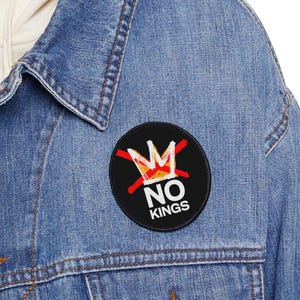May include: A light blue denim jacket with a black circular patch. The patch displays a red crossed-out crown and the white text "NO KINGS". The jacket has a button-down design and pockets.