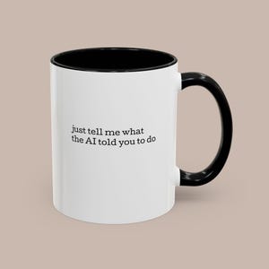 May include: A white ceramic mug with a black interior and handle. The mug features the text "just tell me what the AI told you to do" in a black, sans-serif font. The mug is set against a neutral background.