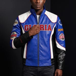 May include: A blue, white, and black leather jacket featuring a stand-up collar, a full-length front zipper, and the word GLORIA in red letters across the chest. The sleeves are adorned with various circular patches, the year 2023, and white script text.