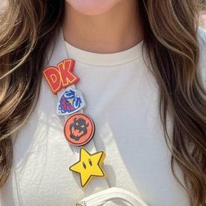 May include: A woman wearing a white t-shirt with a series of colorful character pins. The pins include the letters "DK", a cartoon image, a pumpkin, and a yellow star. A white bag is worn across the body.