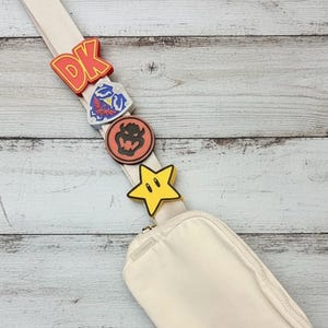May include: A cream-colored crossbody bag with a matching strap, adorned with colorful, cartoon-style charms. The charms include the letters "DK" in red, a blue and white emblem, a brown circular emblem, and a yellow star.