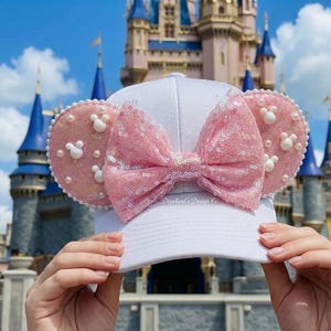May include: A white baseball cap with pink sequined bow and pink mouse ears. The ears are decorated with white pearls and white Mickey Mouse silhouettes. The cap is held up in front of a castle.