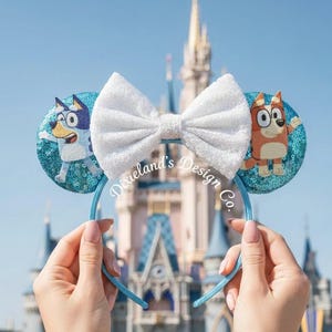 May include: Blue and white sequined mouse ears featuring cartoon characters. The ears have a white bow and are held up in front of a castle. The text "Dixieland's Design Co." is visible.