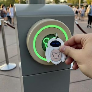 Puede incluir: Un llavero blanco de un personaje de Disney con un icono de Mickey Mouse se sostiene cerca de un punto de acceso gris con un anillo verde brillante. El fondo muestra la entrada a un parque con las palabras "Magic Band".