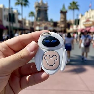 Puede incluir: Llavero blanco y beige con forma de robot, con ojos azules y un logotipo de Mickey Mouse. El llavero se sostiene frente a un fondo borroso de parque temático en un día soleado.