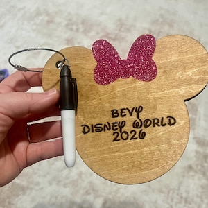 May include: Wooden luggage tag in the shape of Mickey Mouse ears with a glittery pink bow. The tag has the words "Bevy Disney World 2026" engraved on it. A black and white marker is attached to the tag.