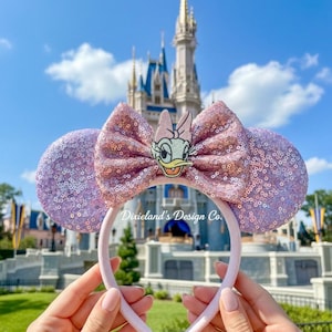 Puede incluir: Unas orejas de ratón con lentejuelas lavanda y un lazo rosa con un diseño de Daisy Duck. La diadema rosa claro se muestra frente a un castillo y un cielo azul. El texto "Dixieland's Design Co." es visible.