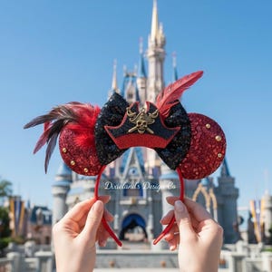 Può includere: Orecchie da topo rosse e nere con paillettes con un design a cappello da pirata. Le orecchie hanno piume rosse e dettagli dorati. La fascia è rossa e il testo "Dixieland's Design Co" è visibile.