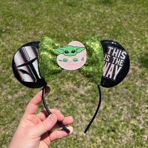 May include: Black headband with two round ears. One ear features a Mandalorian helmet, the other reads "THIS IS THE WAY." A green sequined bow with a Baby Yoda graphic is centered on the headband.