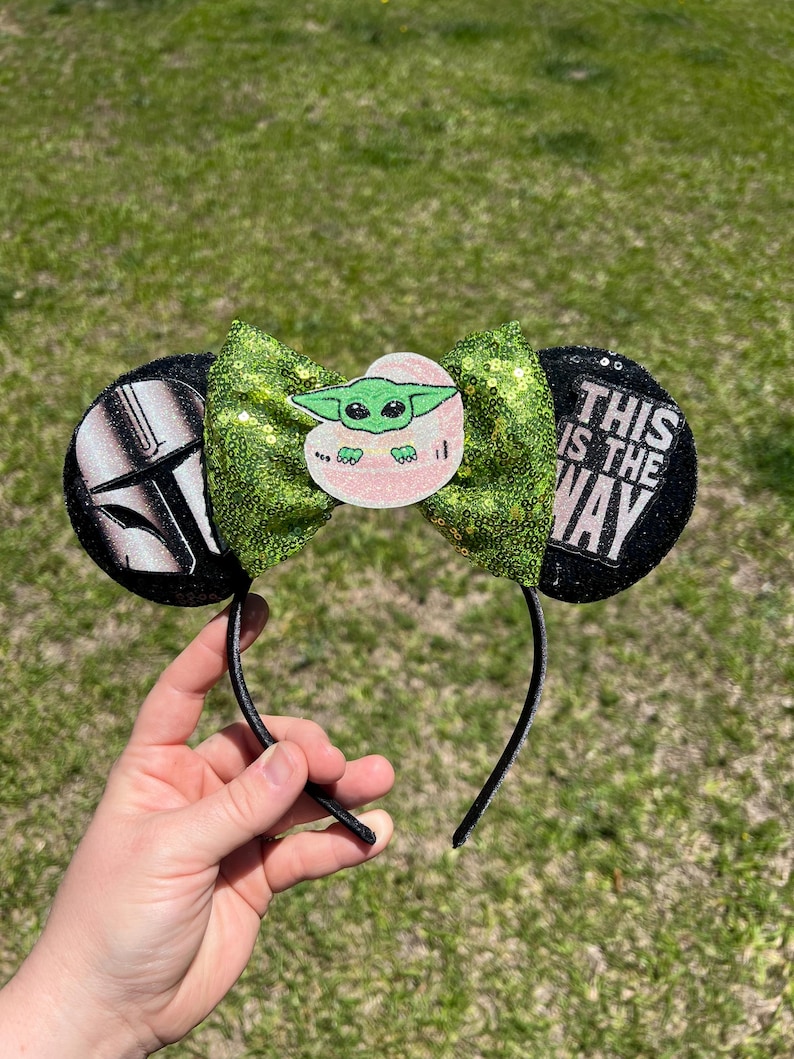 May include: Black headband with two round ears. One ear features a Mandalorian helmet, the other reads "THIS IS THE WAY." A green sequined bow with a Baby Yoda graphic is centered on the headband.