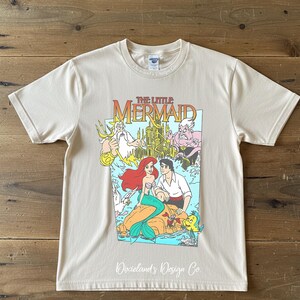 May include: A cream-colored t-shirt featuring a colorful graphic from "The Little Mermaid." The design includes Ariel, Prince Eric, and other characters. The shirt has short sleeves and a crew neck. The text "Dixieland's Design Co." is at the bottom.