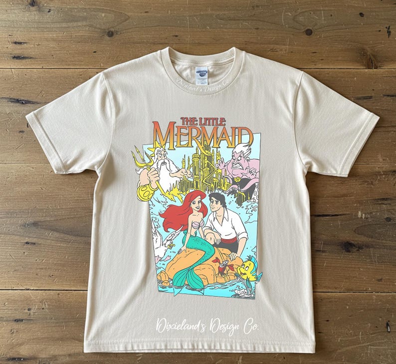 May include: A cream-colored t-shirt featuring a colorful graphic from "The Little Mermaid." The design includes Ariel, Prince Eric, and other characters. The shirt has short sleeves and a crew neck. The text "Dixieland's Design Co." is at the bottom.