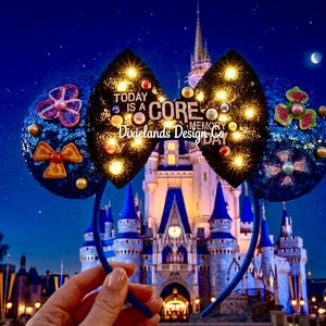 May include: Blue sequined mouse ear headband with a black bow featuring the text "Today is a Core Memory Day". The ears are decorated with colourful embellishments and lights. The headband is held in front of a castle at night.
