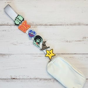 May include: A white fanny pack with a silver strap, adorned with colorful charms. Charms include a green Frankenstein's monster, the letters "DK", a blue shield, a green creature, a witch's hat, and a yellow star.