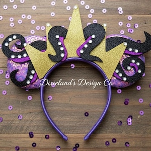 May include: A purple headband with black and gold accents. The headband features two purple sequined ears with black tentacles and gold glitter crowns. The text "Dixieland's Design Co" is visible on the headband.