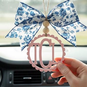 May include: A pink car charm with the letters "ICI" inside a scalloped circle, hanging from a blue floral bow. The bow is white with blue floral patterns. The charm is suspended by a string with wooden beads.