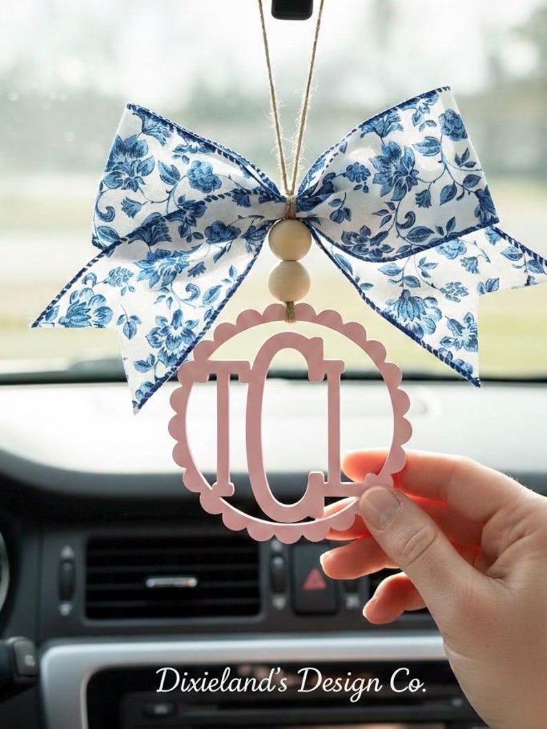 May include: A pink car charm with the letters "ICI" inside a scalloped circle, hanging from a blue floral bow. The bow is white with blue floral patterns. The charm is suspended by a string with wooden beads.