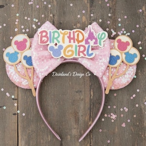 May include: Pink sequined headband with a large bow featuring the text "BIRTHDAY GIRL" and a castle graphic. The headband has two ear-shaped accents with Mickey Mouse balloon designs. The design includes glitter and pastel colors.