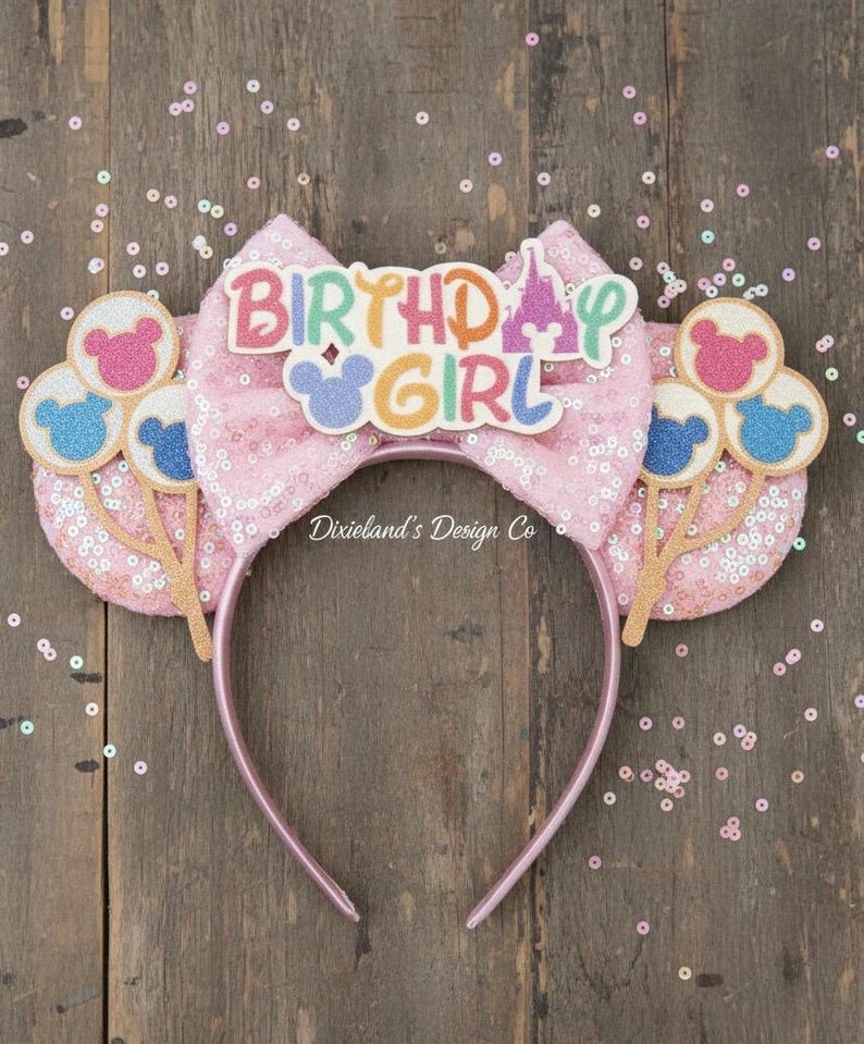 May include: Pink sequined headband with a large bow featuring the text "BIRTHDAY GIRL" and a castle graphic. The headband has two ear-shaped accents with Mickey Mouse balloon designs. The design includes glitter and pastel colors.