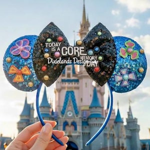 May include: A pair of sequined mouse ears with a blue headband. The ears are blue and black, with colourful embellishments and the text "TODAY IS A CORE MEMORY DAY". The background features a castle.