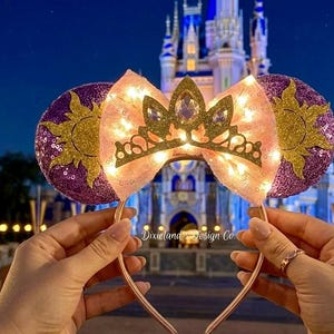 May include: A pair of light-up princess-themed mouse ears. The ears are purple with a gold sun design. The bow is pink with a gold crown and clear gems. The background is a castle at night.