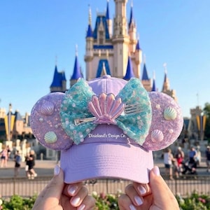 Op de afbeelding: Een lila baseballpet met muisoren en een schelpenstrik. De oren zijn versierd met schelpen en pailletten. De strik is turquoise en roze. De tekst "Dixieland's Design Co." staat op de pet.