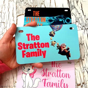 以下が含まれることがあります： 赤色の「The Stratton Family」という文字が入ったナンバープレート。1枚は風船で浮いている家の漫画の画像、もう1枚は猫、3枚目は未来的なオートバイです。