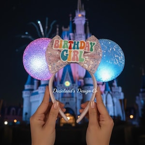Op de afbeelding: Een roze haarband met pailletten oren en een strik met de tekst "BIRTHDAY GIRL" in kleurrijke letters. De oren lichten op met paarse en blauwe tinten. De achtergrond toont een kasteel en vuurwerk.