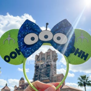 May include: A lime green headband with two round ears. The ears have the text "OOH" and "HH" on them. A blue sequined bow with three white eyes is in the center. A silver claw graphic is on each ear.