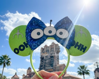 May include: A lime green headband with two round ears. The ears have the text "OOH" and "HH" on them. A blue sequined bow with three white eyes is in the center. A silver claw graphic is on each ear.