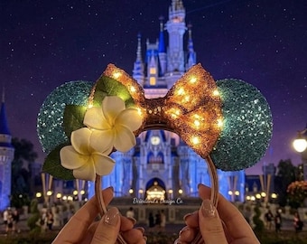 May include: A pair of mouse ears with a sequined gold bow and teal sequined ears. The ears are decorated with white flowers and green leaves. The background features a castle at night.
