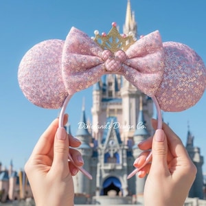 Pode incluir: Tiara de orelhas de rato rosa com lantejoulas com laço a condizer e coroa dourada. As orelhas são seguradas contra um céu azul com um castelo ao fundo. O laço apresenta um emblema do Mickey Mouse.