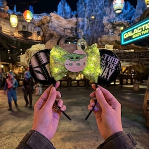 May include: Black mouse ears with a green sequined bow featuring a Baby Yoda character and the phrase "This is the Way." One ear has a Mandalorian helmet design. The ears are held up in front of a Star Wars-themed background with lanterns.