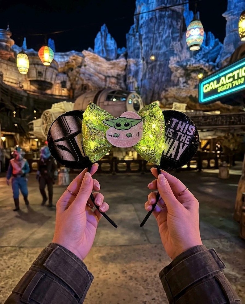 May include: Black mouse ears with a green sequined bow featuring a Baby Yoda character and the phrase "This is the Way." One ear has a Mandalorian helmet design. The ears are held up in front of a Star Wars-themed background with lanterns.