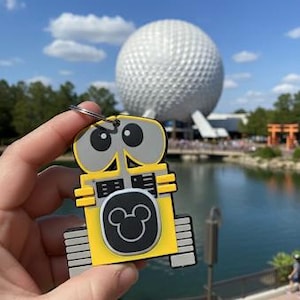 May include: A yellow and gray WALL-E keychain with a black Mickey Mouse emblem. The keychain is held in front of a large white sphere and a body of water. The background includes trees, a bridge, and a blue sky with clouds.