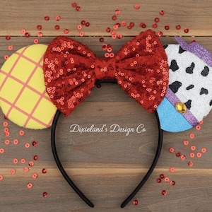 May include: A black headband with character-themed ears. The ears are yellow with a pink grid pattern, white with black spots, and blue. A red sequined bow is centered on the headband. The text "Dixieland's Design Co" is visible.