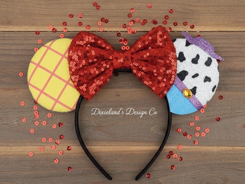 May include: A black headband with character-themed ears. The ears are yellow with a pink grid pattern, white with black spots, and blue. A red sequined bow is centered on the headband. The text "Dixieland's Design Co" is visible.