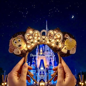 May include: Glittering brown and gold mouse ears with a tiara design and lights, held in front of a castle at night. One ear features a cartoon dog, the other a golden apple. The text "Dixielands Design Co" is visible.