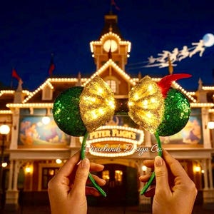 Pode incluir: Dois arcos de orelhas com o tema Peter Pan. As orelhas são verdes com lantejoulas e os laços são dourados com lantejoulas, iluminados com luzes brancas quentes. Uma orelha tem uma pena vermelha. O fundo mostra um edifício e luzes à noite.