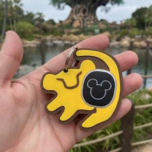Op de afbeelding: Gele en bruine sleutelhanger met een leeuwenwelp-ontwerp, een zwart vierkant en een Mickey Mouse-silhouet. De sleutelhanger wordt vastgehouden voor een vage achtergrond van een park met een grote boom en water.