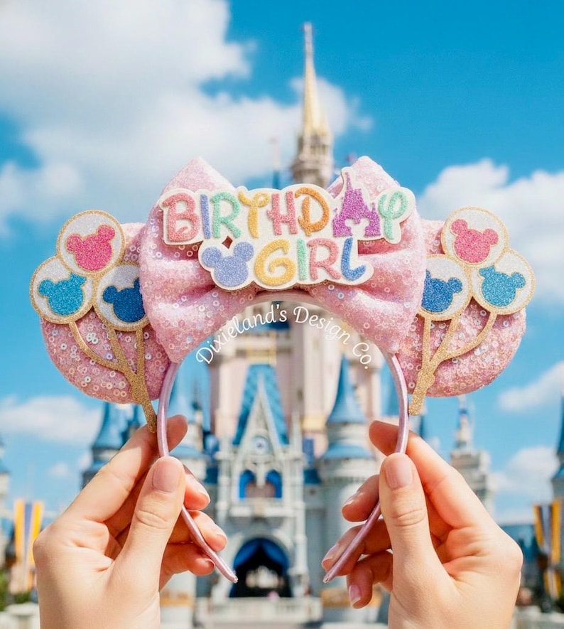 May include: Pink sequined headband with a large bow featuring the text "BIRTHDAY GIRL". The headband has Mickey Mouse balloon accents and is set against a backdrop of a castle under a blue sky.
