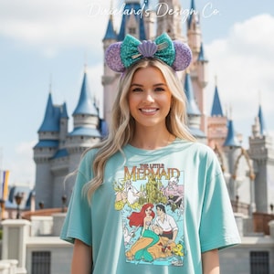 May include: A light blue t-shirt featuring a graphic from "The Little Mermaid" with Ariel and Prince Eric. The model is wearing a pair of purple and teal sequined mouse ears with a seashell and fork detail. The background features a castle.