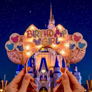May include: Pink sequined headband with the words "BIRTHDAY GIRL" in colorful letters. The headband has Mickey Mouse ears and balloon accents. The background features a castle at night.