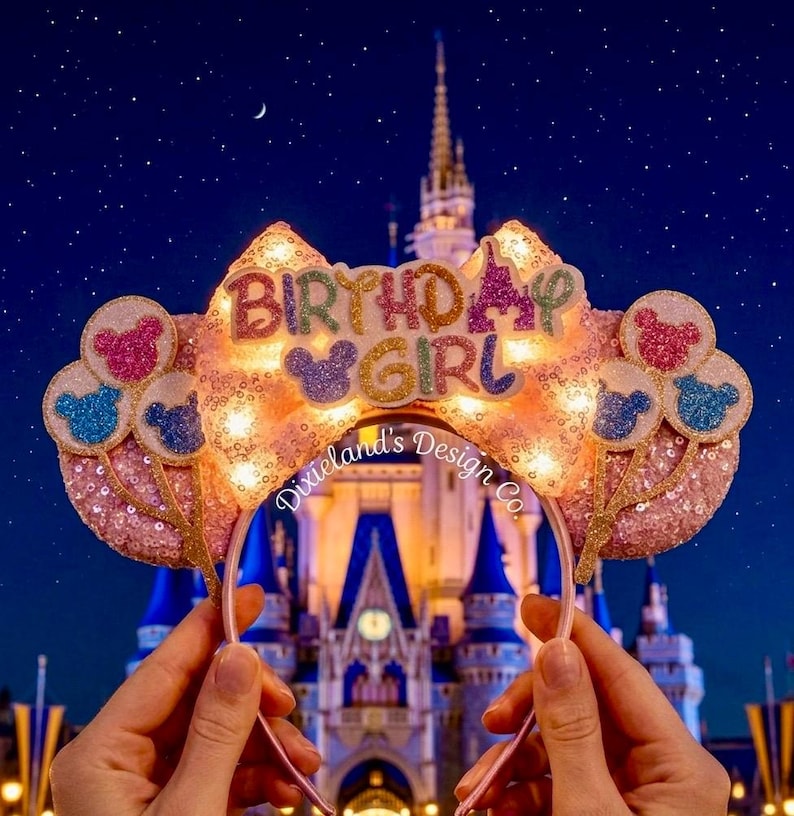 May include: Pink sequined headband with the words "BIRTHDAY GIRL" in colorful letters. The headband has Mickey Mouse ears and balloon accents. The background features a castle at night.